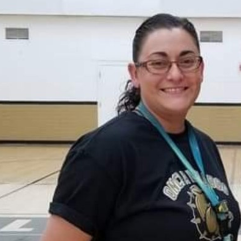 A photo of a white woman with brown hair in a pony tail. She is wearing glasses and a school t-shirt. This is Alexandra Bustillos.