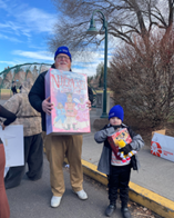 A man and a boy, both wearing blue school caps, stand next to each other holding up their homemade creations at the 2026 Bend Christmas Parade. 