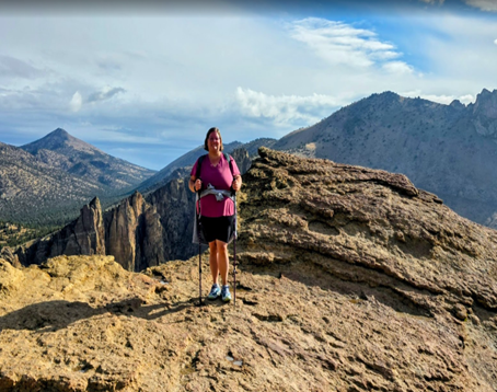 A woman wearing black shorts and a pink t-shirt and holding a walking stick while posing at the top of a mountain peak, with other mountains behind her.